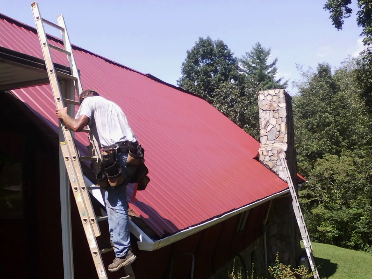Roofer on a ladder beside a red metal roof and stone chimney — Soffit & Fascia Repair by Roofing Brothers in Boling